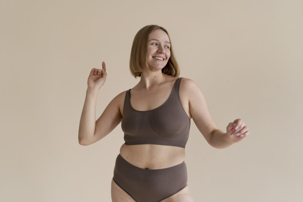 Smiling woman in brown underwear dancing playfully against neutral beige background.