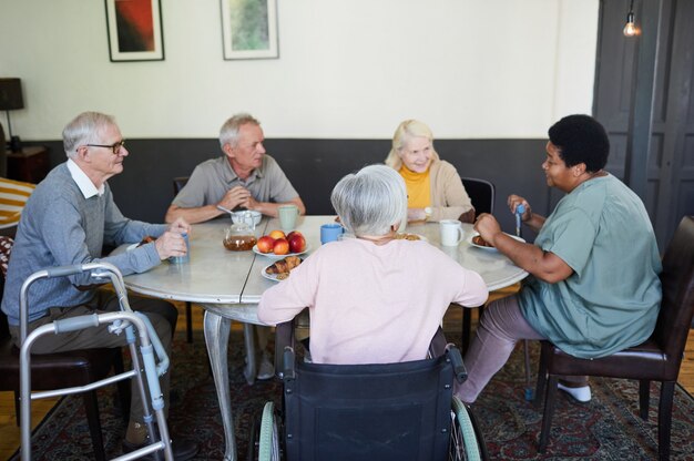 Group of seniors sitting around a table, chatting and enjoying tea and fruit in a cozy indoor setting.