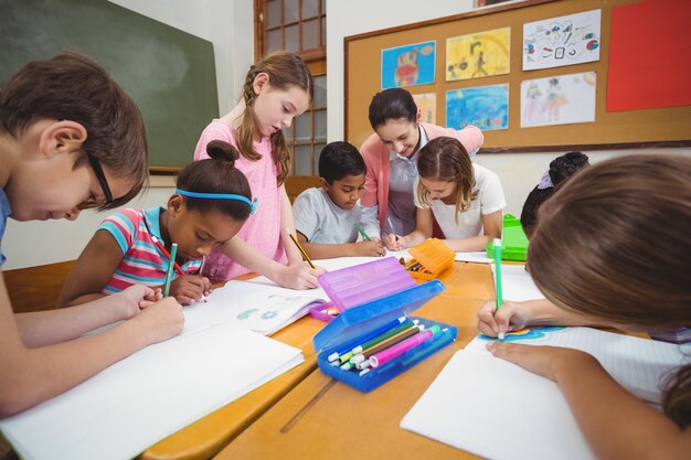A group of children gathered around a classroom table drawing pictures with markers and pencils while a teacher helps them.
