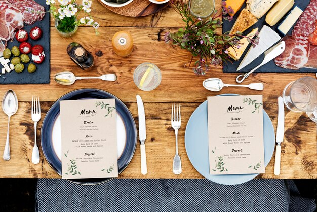 Styled dining table with menu cards, plates, silverware, and a variety of cheeses, meats, and appetizers on wooden boards.