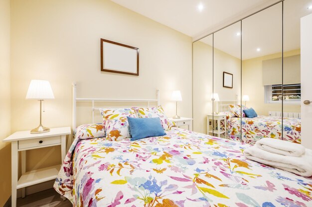 Cozy bedroom with floral bedspread, white bedside tables, lamps, and mirrored wardrobe reflecting the room.