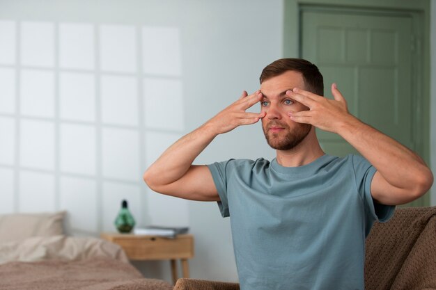 Man gently massaging under-eye area while sitting on a couch in a bright, modern living room.