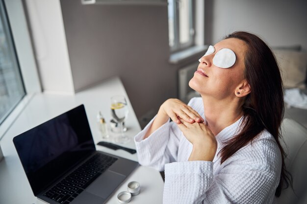 Woman relaxing with eye patches on, sitting by a laptop near a window in a calm home setting.