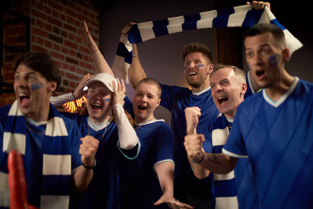 Group of fans in blue jerseys celebrating indoors with scarves and raised fists during a game.

